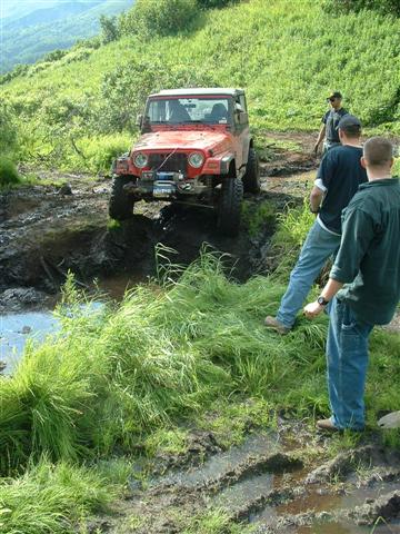 2000 Jeep Wrangler - A red 2000 Jeep TJ navigates a challenging muddy off-road trail with onlookers present.