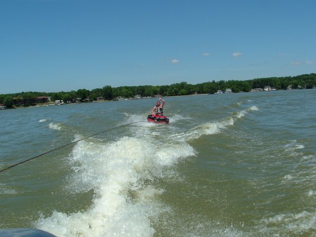 Person riding a red towable tube behind a boat's wake on a lake with wooded shoreline
