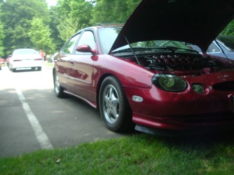 1993 Ford Taurus - This red sedan has its hood up, revealing the engine compartment. A white car is parked nearby on the road.