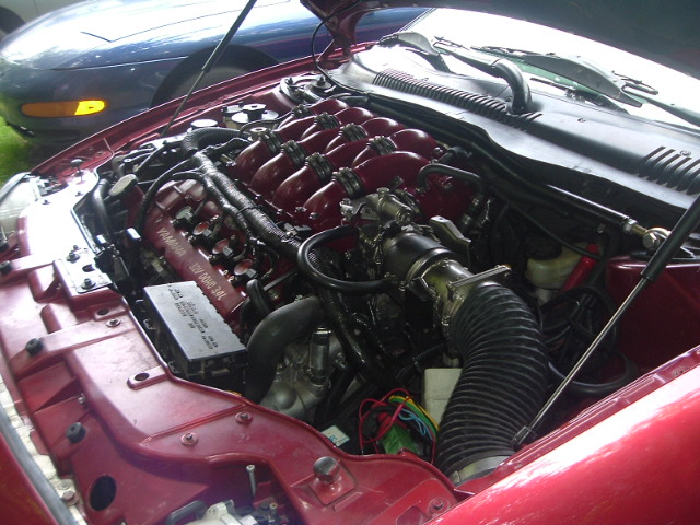 Engine bay of a red car showing a V6 engine with red intake manifolds. A blue car is visible in the background