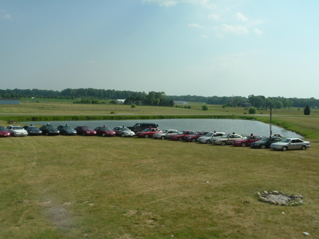 Many cars parked in a grassy field next to a lake under a partly cloudy sky