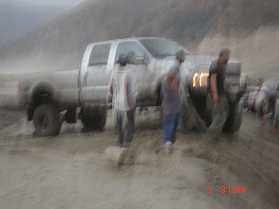 Blurry side view of a muddy silver crew-cab pickup truck with people standing nearby in a mud field