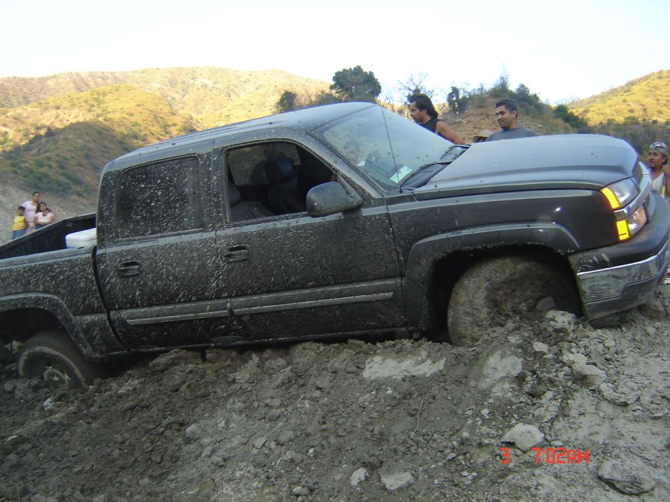 Mud-caked dark gray crew cab pickup climbing a rutted dirt bank at a mud run
