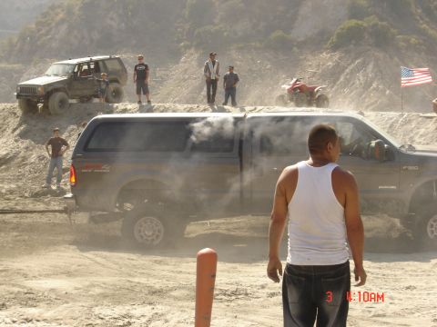 1977 Toyota Land Cruiser - A pickup truck with a camper shell is seen in a dusty, off-road environment with other vehicles and people present.