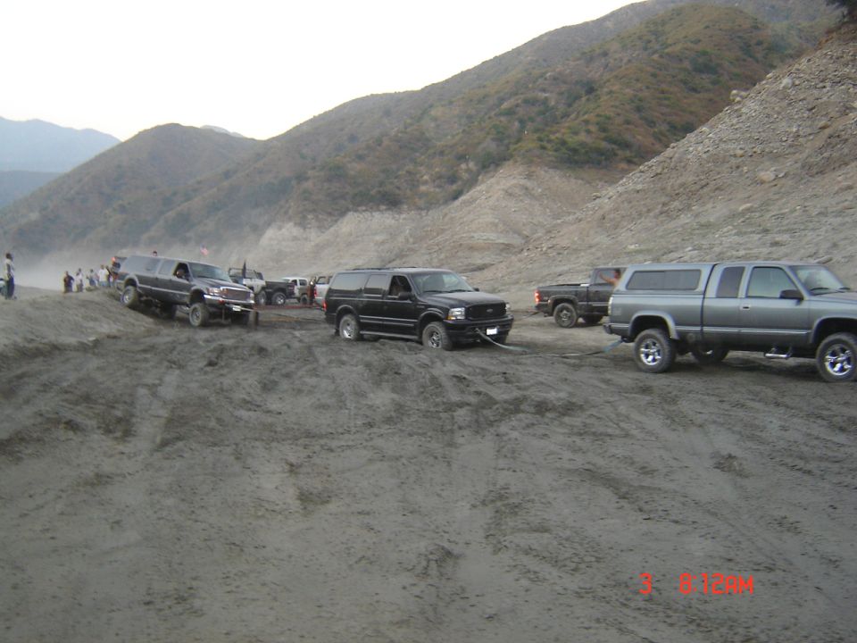 Black Ford Excursion being towed between two pickups in a dusty desert wash