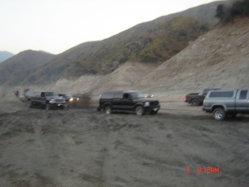 Blurry photo of several trucks and SUVs lined up on a dirt off-road trail below a hillside.