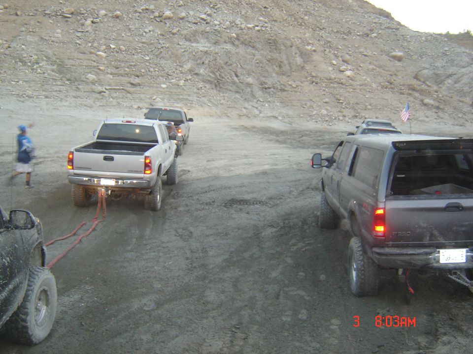 White Chevy pickup pulling a vehicle with tow straps up a muddy dirt hill, gray Ford truck at right