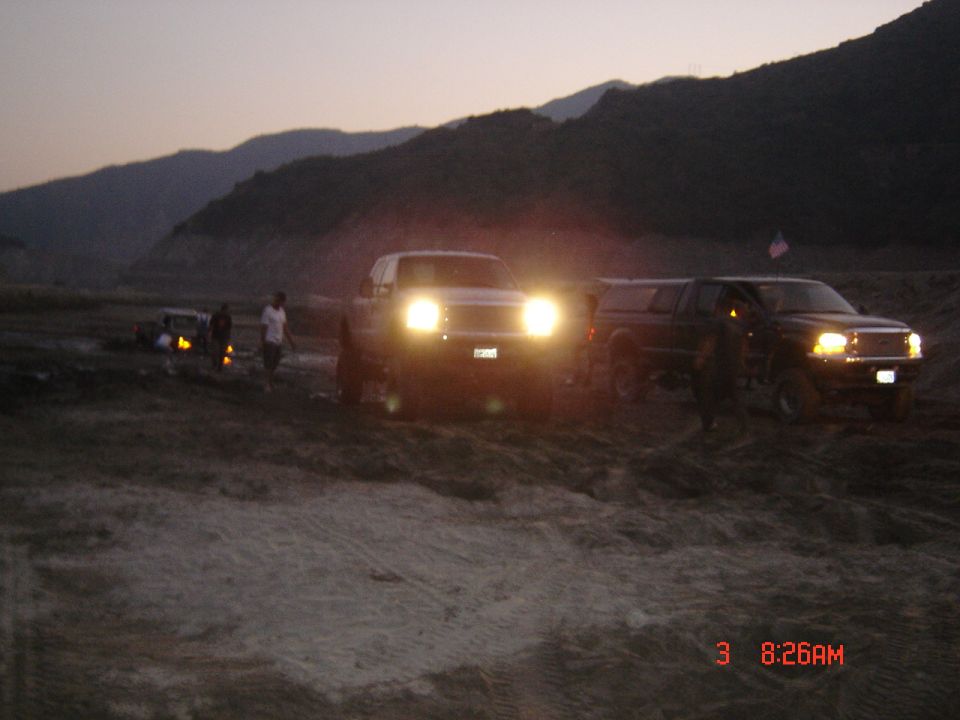 Two lifted trucks with headlights on in a sandy off-road area at dusk, mountains behind