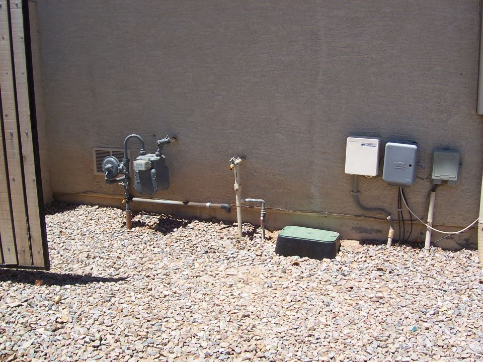 Gas meter, pipes, and utility boxes on a stucco house wall over a gravel side yard