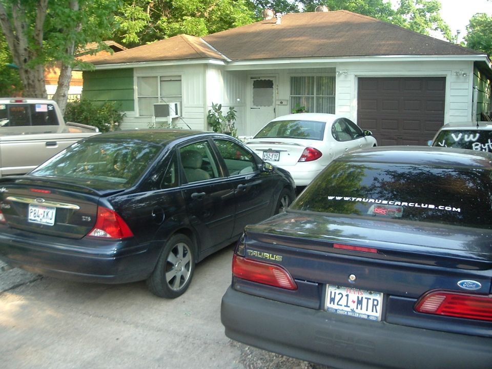 Dark blue Ford Taurus with a sticker on the back window, parked next to a black Ford Focus and a white car. A pickup truck is also visible