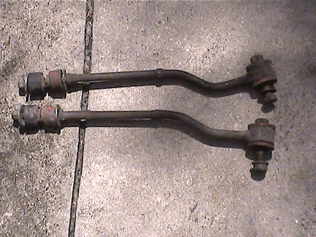 Two dark metal suspension control arms with rubber bushings and bolts, resting on a concrete surface