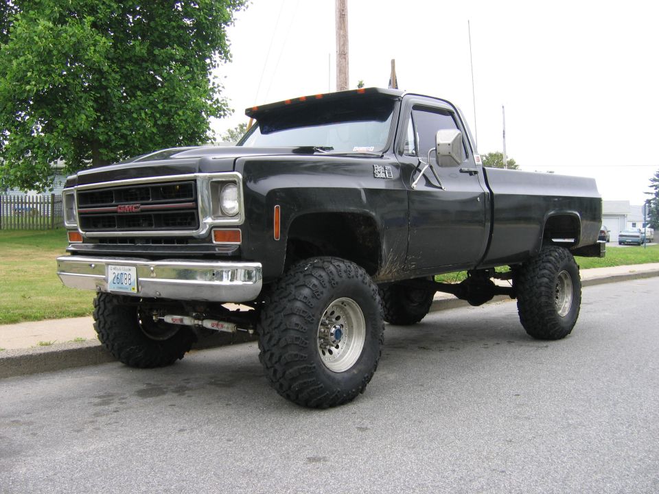 Black lifted GMC pickup on big mud tires parked at a curb, front three-quarter view