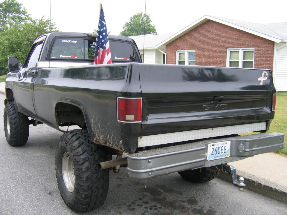 Rear of black lifted GMC pickup with American flag in bed, parked at curb by brick house