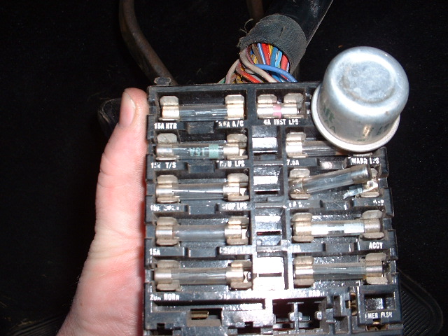 Hand holding an automotive fuse panel with glass fuses, labels and a silver flasher can