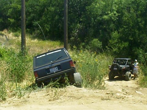 1986 Ford Bronco - Two off-road vehicles navigate a dirt path surrounded by greenery, with one Jeep tilted on its side.