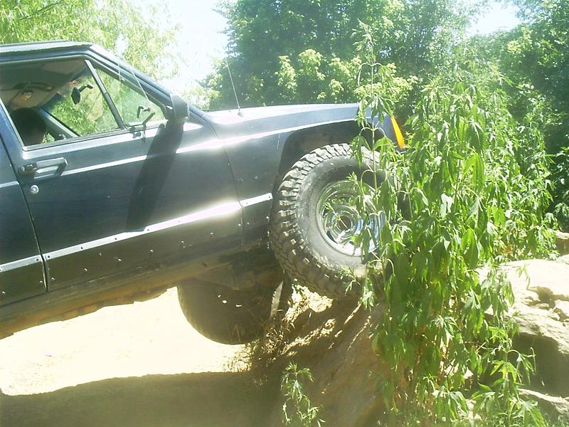 Black Jeep Cherokee XJ with off-road tires climbing over rocks and brush