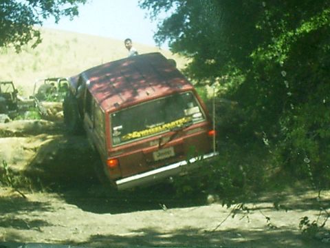 1986 Ford Bronco - A red Jeep Cherokee navigates a rocky, off-road trail with another vehicle visible in the background.