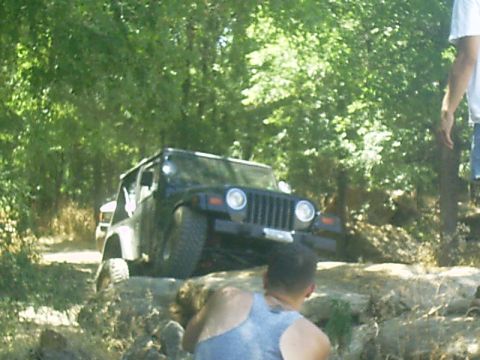 1986 Ford Bronco - A black Jeep Wrangler TJ navigates a rocky off-road path with trees in the background.