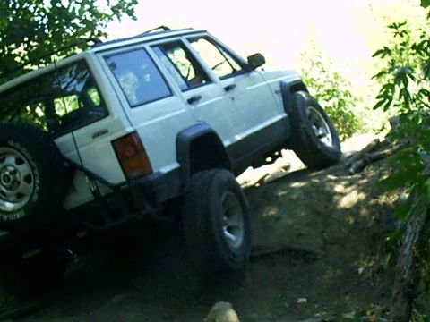 1986 Ford Bronco - A white Jeep Cherokee XJ navigates a challenging off-road obstacle course, climbing over fallen logs and uneven terrain.