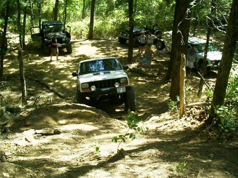 1986 Ford Bronco - Several off-road vehicles are gathered in a wooded area, with one Jeep navigating a dirt mound.