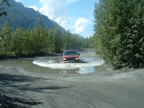 1994 Ford E350 - A red van navigates a large puddle on a dirt road, surrounded by trees and mountains under a partly cloudy sky.