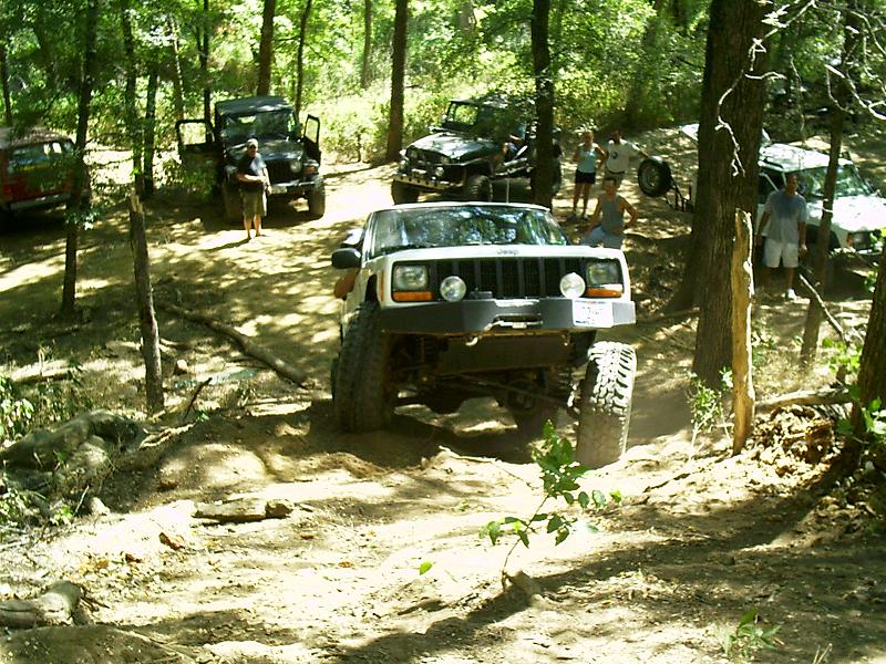 Modified Jeep Cherokee XJ with large tires on a dirt trail, other Jeeps and people in background