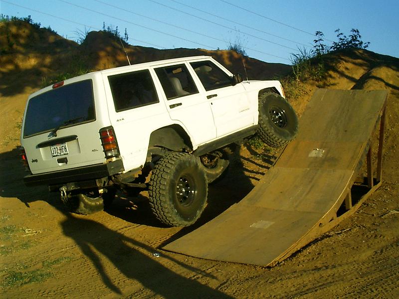 White Jeep Cherokee XJ with large tires climbing a wooden ramp