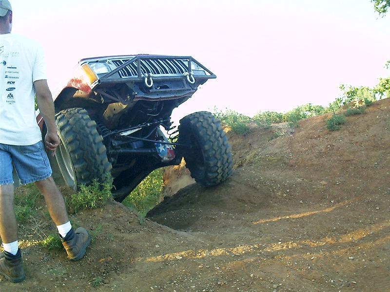 off-road vehicle with large tires climbing a steep dirt hill, person standing nearby