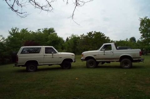 1985 Ford Bronco - Two white Ford Broncos, an SUV and a pickup truck, are parked side-by-side on a grassy field under a cloudy sky.