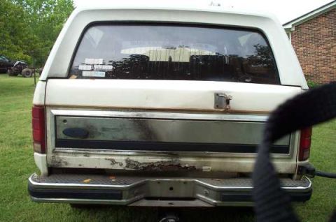 1985 Ford Bronco - The rear of a white Ford Bronco, with visible rust on the lower tailgate and bumper.