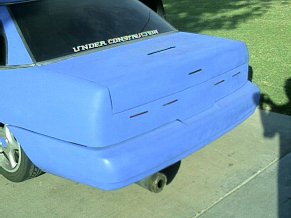 Rear of primer-blue sedan with 'UNDER CONSTRUCTION' decal on rear window, parked on driveway.