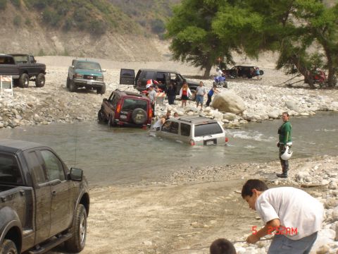 1977 Toyota Land Cruiser - Multiple off-road vehicles navigate a shallow, rocky river, with onlookers observing the scene.