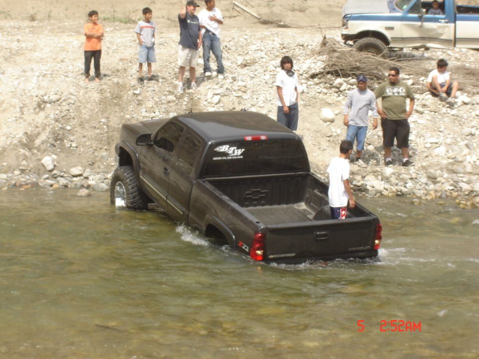 Lifted black Chevrolet Silverado extended cab backed into a shallow river with onlookers
