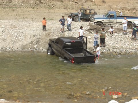 1977 Toyota Land Cruiser - Several people watch as a black pickup truck drives through a rocky riverbed with other vehicles nearby.