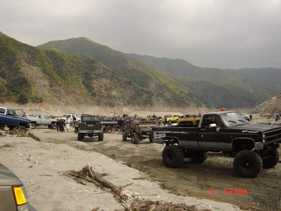 Several lifted pickup trucks and SUVs parked in a sandy, mountainous area. One black Dodge pickup is prominent in the foreground