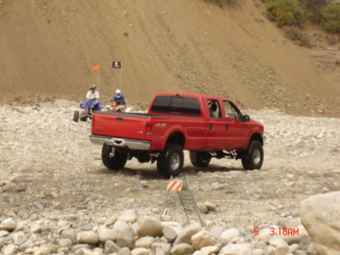 1977 Toyota Land Cruiser - A red pickup truck navigates a rocky, off-road environment with ATVs in the distance.