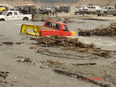 1977 Toyota Land Cruiser - Multiple vehicles are seen in a riverbed, with one red and yellow pickup truck partially submerged in debris-filled water.