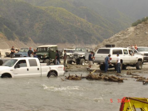 1977 Toyota Land Cruiser - A group of off-road vehicles navigate through a shallow body of water with a mountainous backdrop.