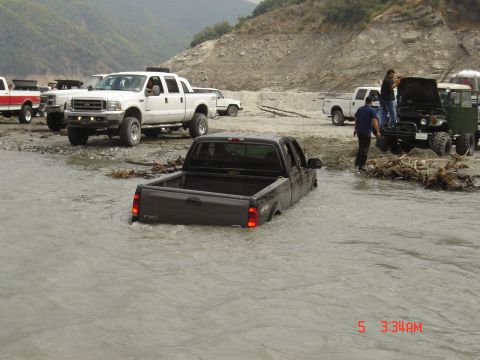1977 Toyota Land Cruiser - Several trucks are stranded in a river, with one dark gray pickup truck submerged up to its doors.