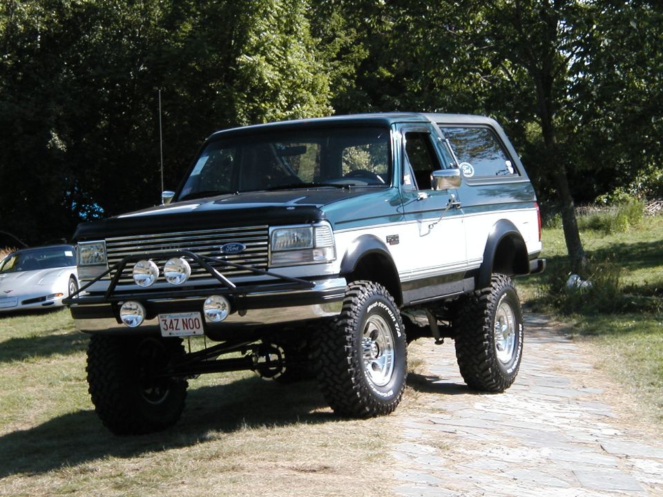 Lifted 1994 Ford Bronco, green and white two-tone paint, aftermarket grille guard with four lights, off-road tires. White sports car visible in background
