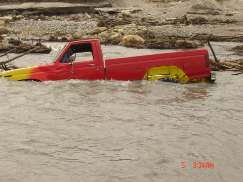 1977 Toyota Land Cruiser - A pickup truck is stuck in floodwaters, with debris and rocks visible on the riverbank.