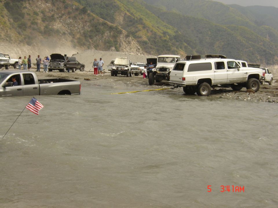 Silver Dodge Ram bed-deep in muddy river with recovery strap to a lifted white Suburban