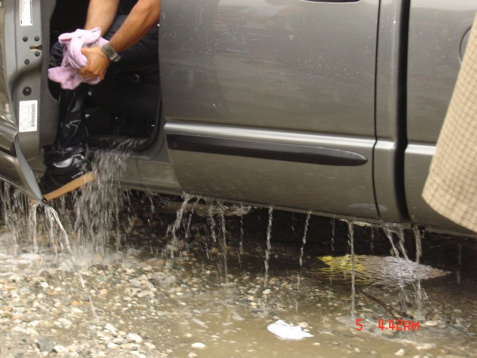 Water draining from the open door and rocker of a flooded gray pickup on a gravel bar