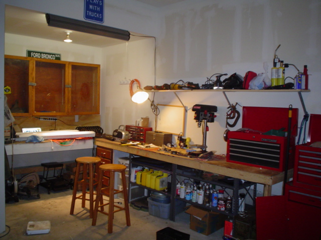 Home garage workshop with workbench, drill press, red Craftsman tool chest, two wooden stools, and Ford Bronco sign