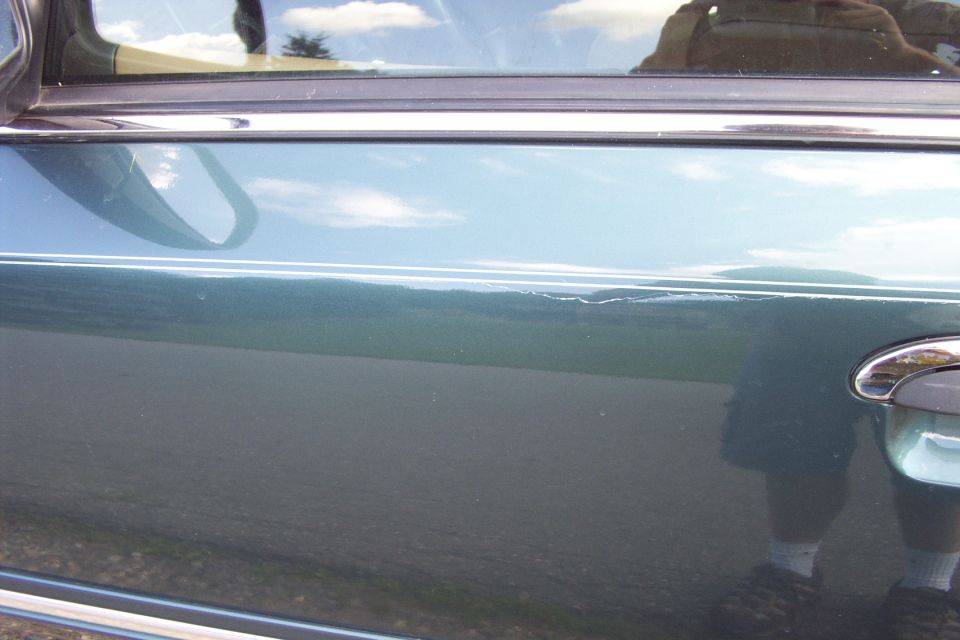 Close-up of a dark green car door with chrome trim and door handle, scratch visible, people reflected