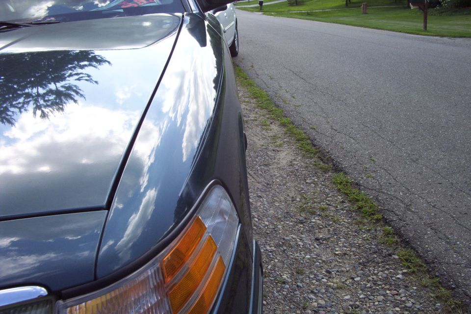 Close-up of dark green sedan's hood and amber corner light, parked on gravel shoulder of a residential road