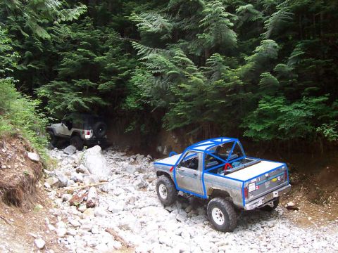 1984 Ford Bronco II - Two modified trucks tackle a challenging, rocky path through a dense forest.