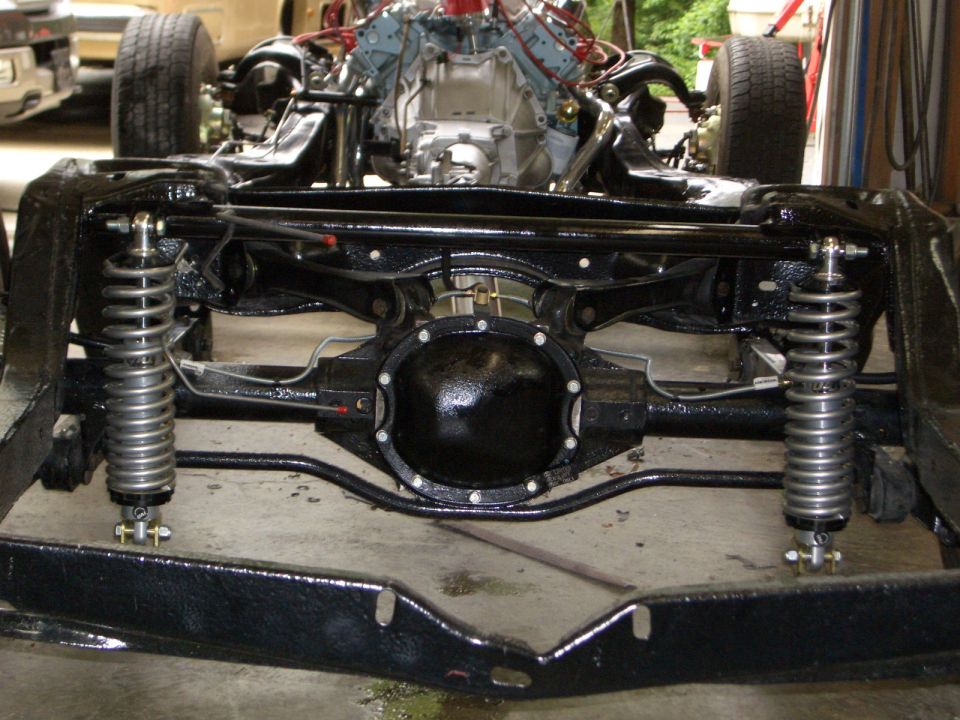 Bare black car frame with rear axle, coilover shocks and mounted V8 in a shop