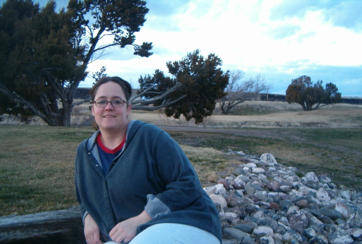 Person in a gray hooded sweatshirt and glasses kneeling on a lawn near a rock border at dusk