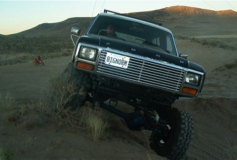 1986 Ford Bronco - This lifted Ford Bronco navigates a sandy incline, with a dirt bike rider visible in the background.